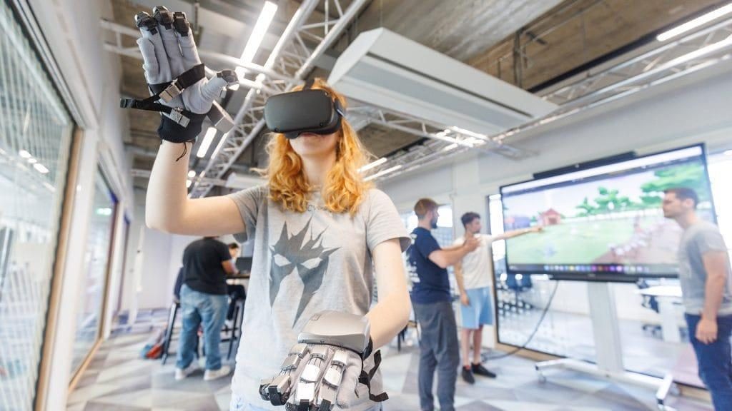 Een studente met een VR-bril en haptische handschoenen grijpt in de lucht; achter haar overleggen studenten bij een scherm met een virtuele wereld.